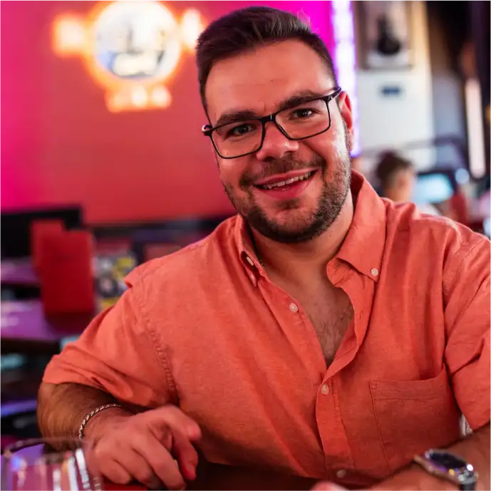Marco Rodrigues coach. Adulto, tem 32 anos. Ele está usando uma camisa social de botões, aberta no primeiro botão, ele usa óculos, barba serrada por fazer, cabelos curto. Ele está sorrindo e olhando para a câmera em um ambiente de bar com muitas luzes desfocadas ao fundo.